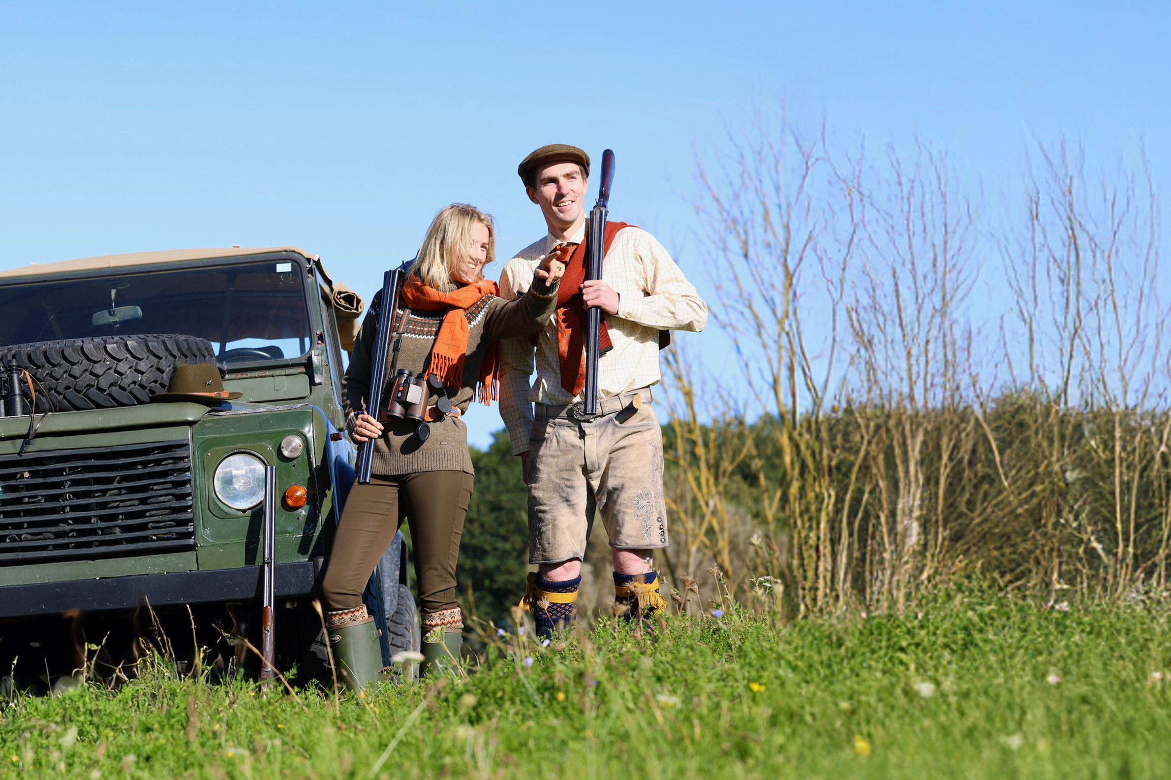 Eine Frau und ein Mann in Jagdkleidung stehen mit Flinten neben einem grünen Geländewagen auf einer Wiese, die im Hintergrund in einen grünen Wald übergeht. Die Frau zeigt in die Ferne, beide blicken in dieselbe Richtung.