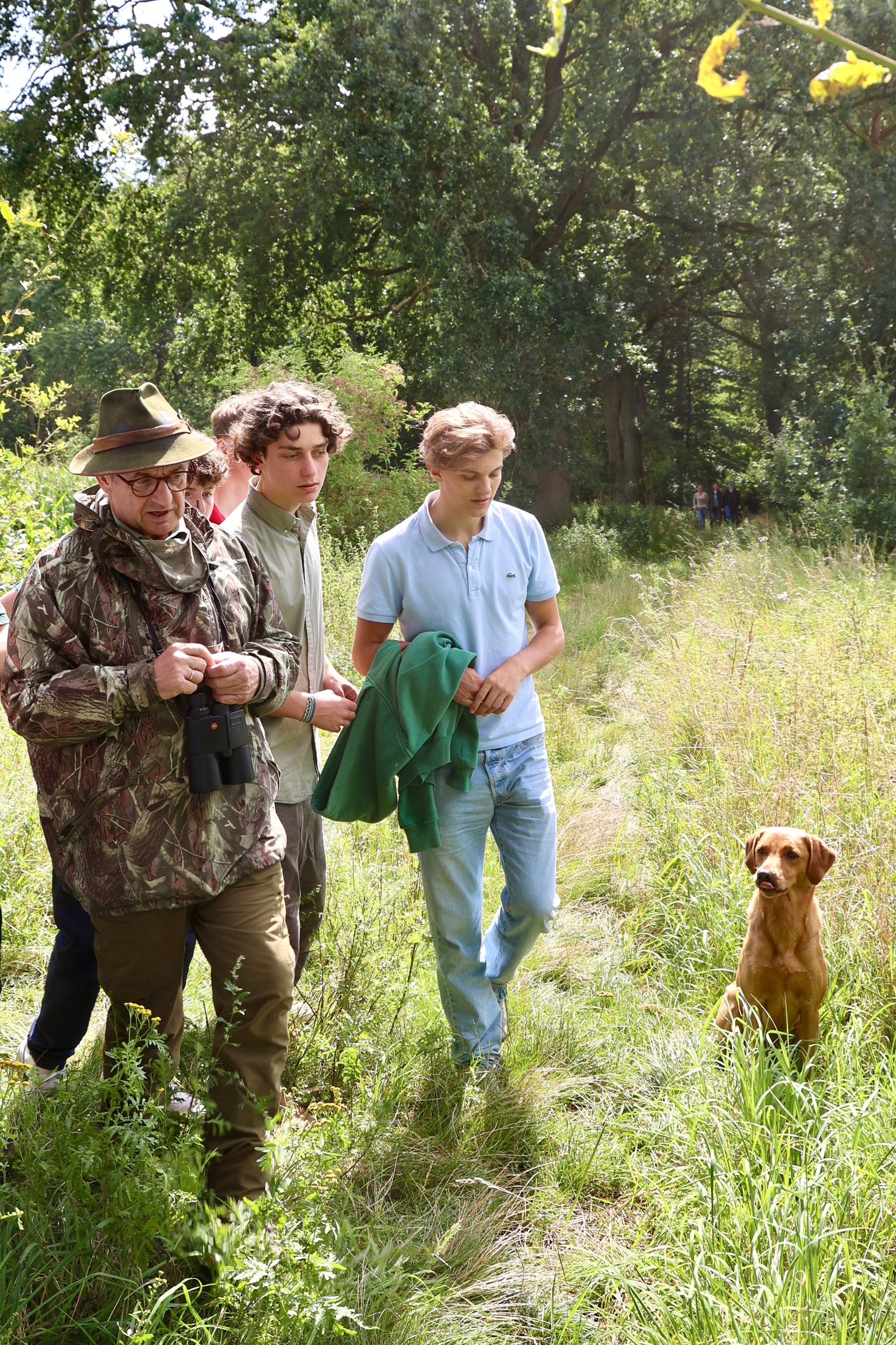 Eine Gruppe junger Männer wird von einem Jagdausbilder in Tarnbekleidung mit Fernglas auf einem Wiesenpfad im Vordergrund eines grün belaubten Waldes begleitet. Am Wegesrand sitzt ein aufmerksamer Jagdhund.