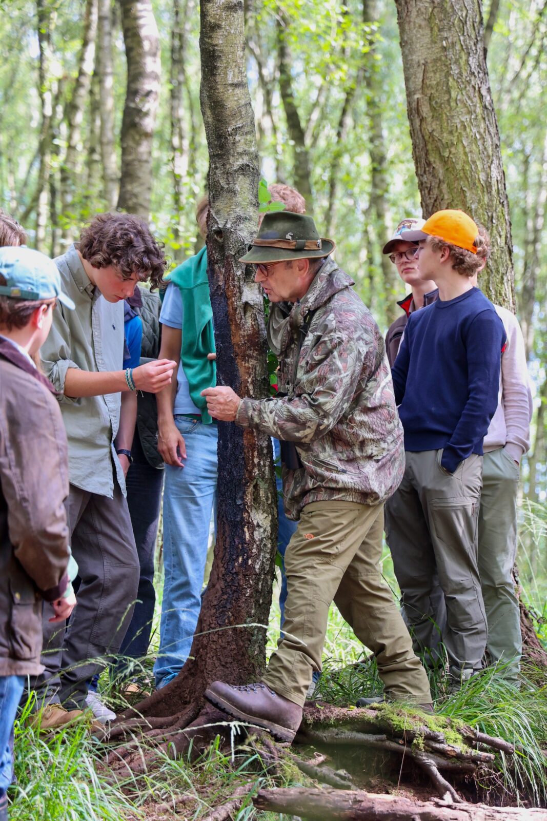 Ein Jagdausbilder in Tarnkleidung erklärt einer Gruppe junger Menschen an einem Baumstamm Details zu einer Wildspur. Die Gruppe steht im Halbkreis um den Baum, im Hintergrund ein lichter Wald mit grün belaubten, schlanken Bäumen.