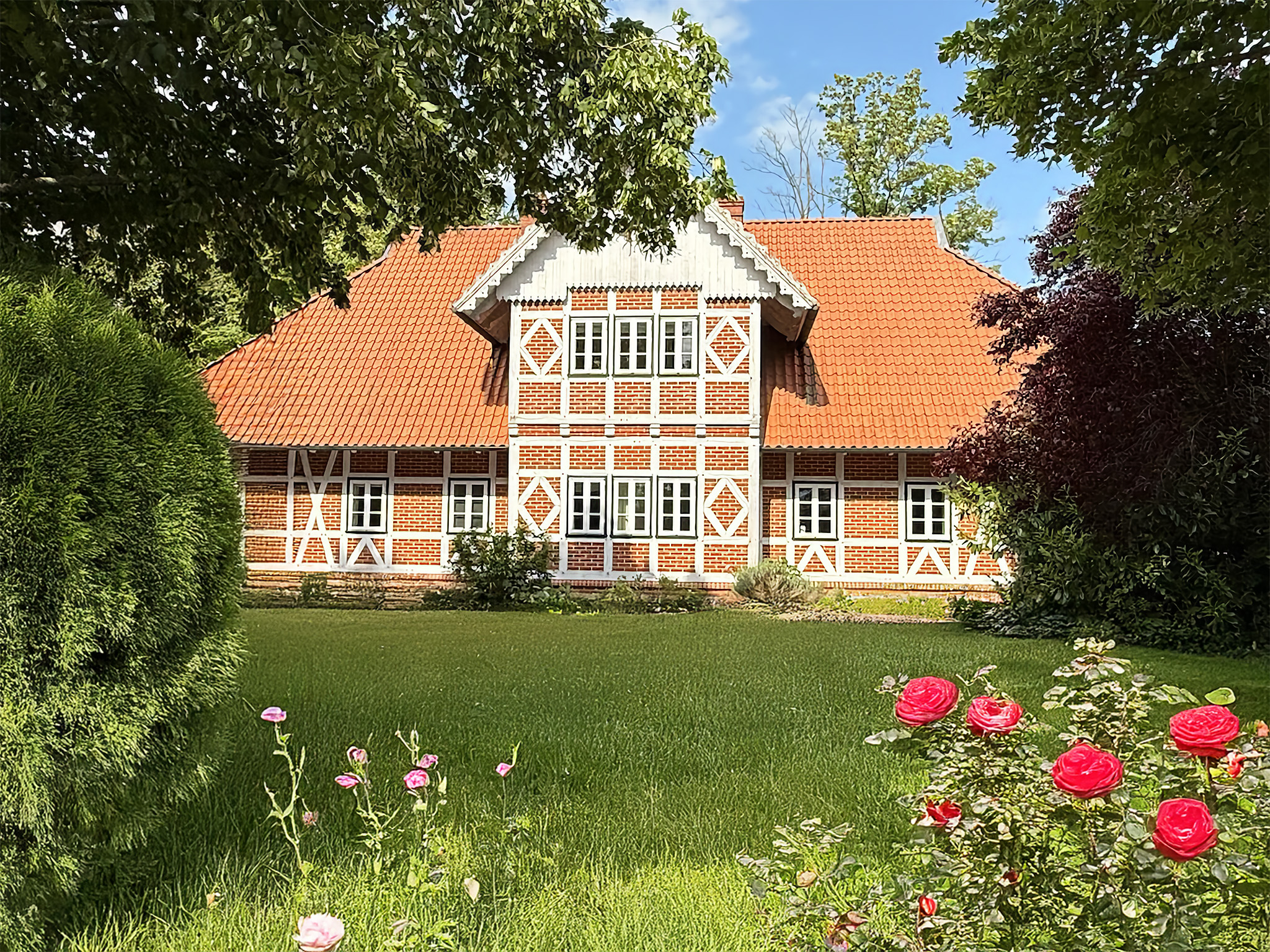 Fachwerkhaus mit rotem Ziegeldach und rotbraunen Ziegelsteinen inmitten eines gepflegten, grünen Gartens mit blühenden Rosen sowie umliegenden Bäumen und Büschen bei Sonnenschein.