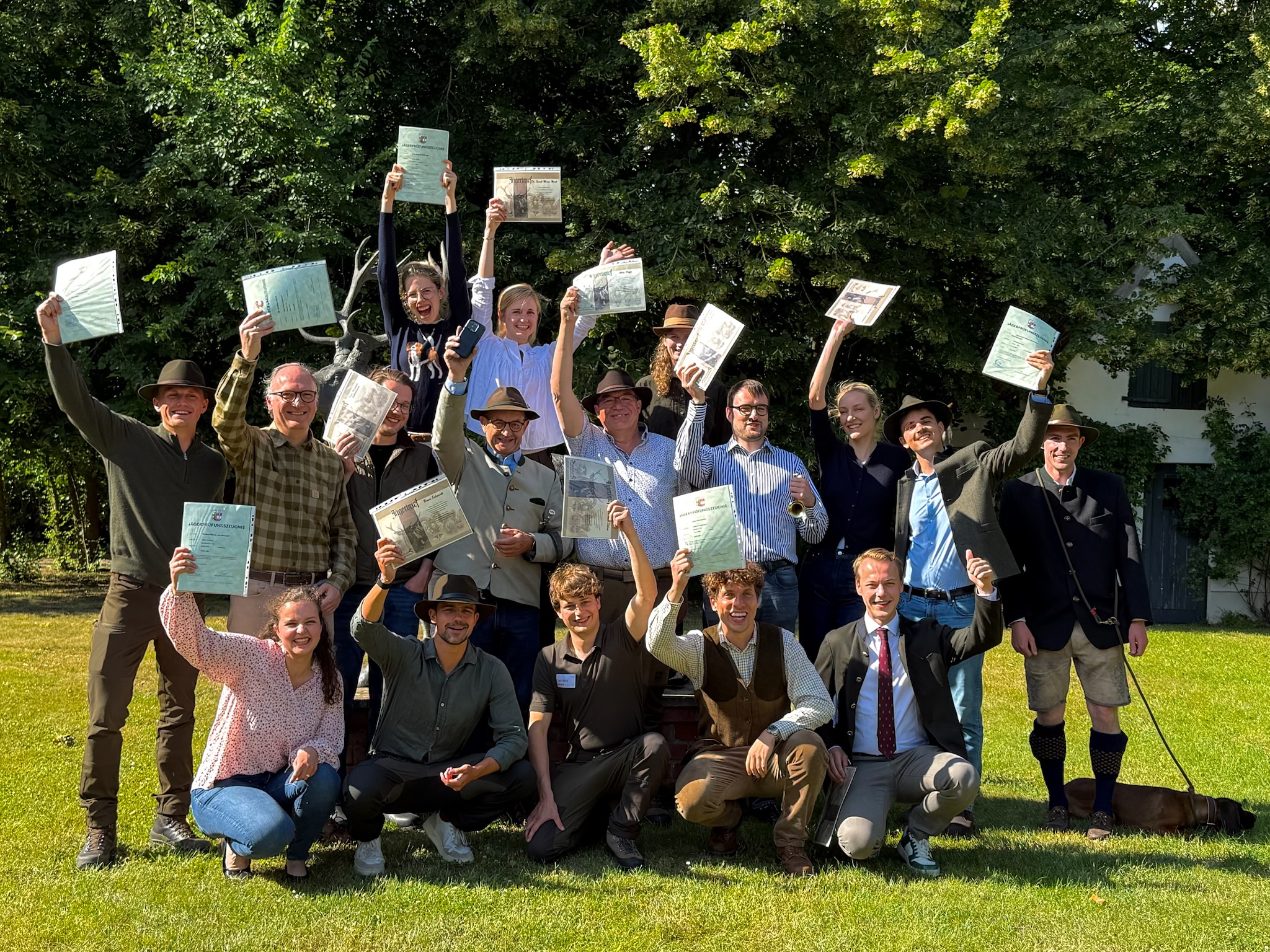 Eine Gruppe fröhlich lächelnder Absolventinnen und Absolventen steht und kniet auf einer sonnenbeschienenen Wiese - viele halten ihre Jagdscheine jubelnd in die Höhe. Im Hintergrund stehen eine Hirsch-Statue, grüne Bäume und ein helles Gebäude mit grünen Fensterläden.