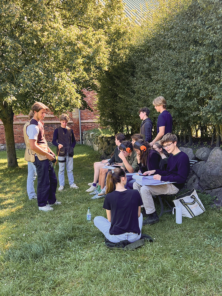 Eine Gruppe junger Erwachsener sitzt auf einer Bank auf einer Wiese, während einige weitere daneben stehen. Die Sitzenden schauen konzentriert in ihre Mappen - zwei von ihnen tragen Gehörschutz. Im Hintergrund steht ein großer Baum und eine Steinmauer bepflanzt mit hohen Büschen.