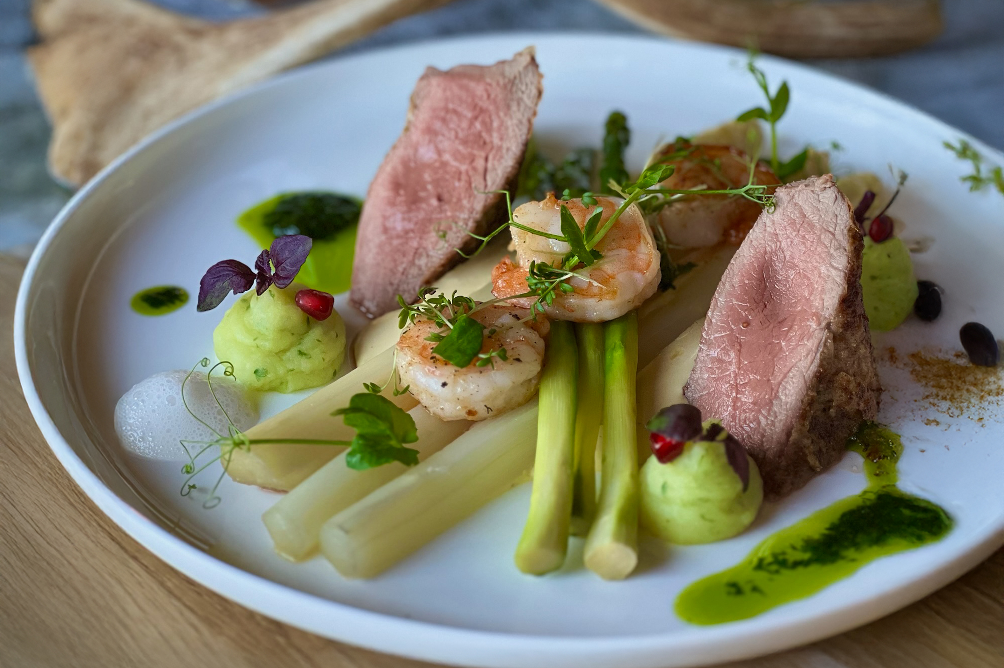 Aufwendig und detailreich angerichtetes Gericht mit Wildfleisch, Spargel, Garnelen, Espuma, Kartoffelpüree und wilden Kräutern, kunstvoll auf einem weißen Teller drapiert.
